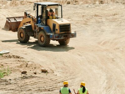 Construction workers in safety gear walking at a busy site with a backhoe in operation.